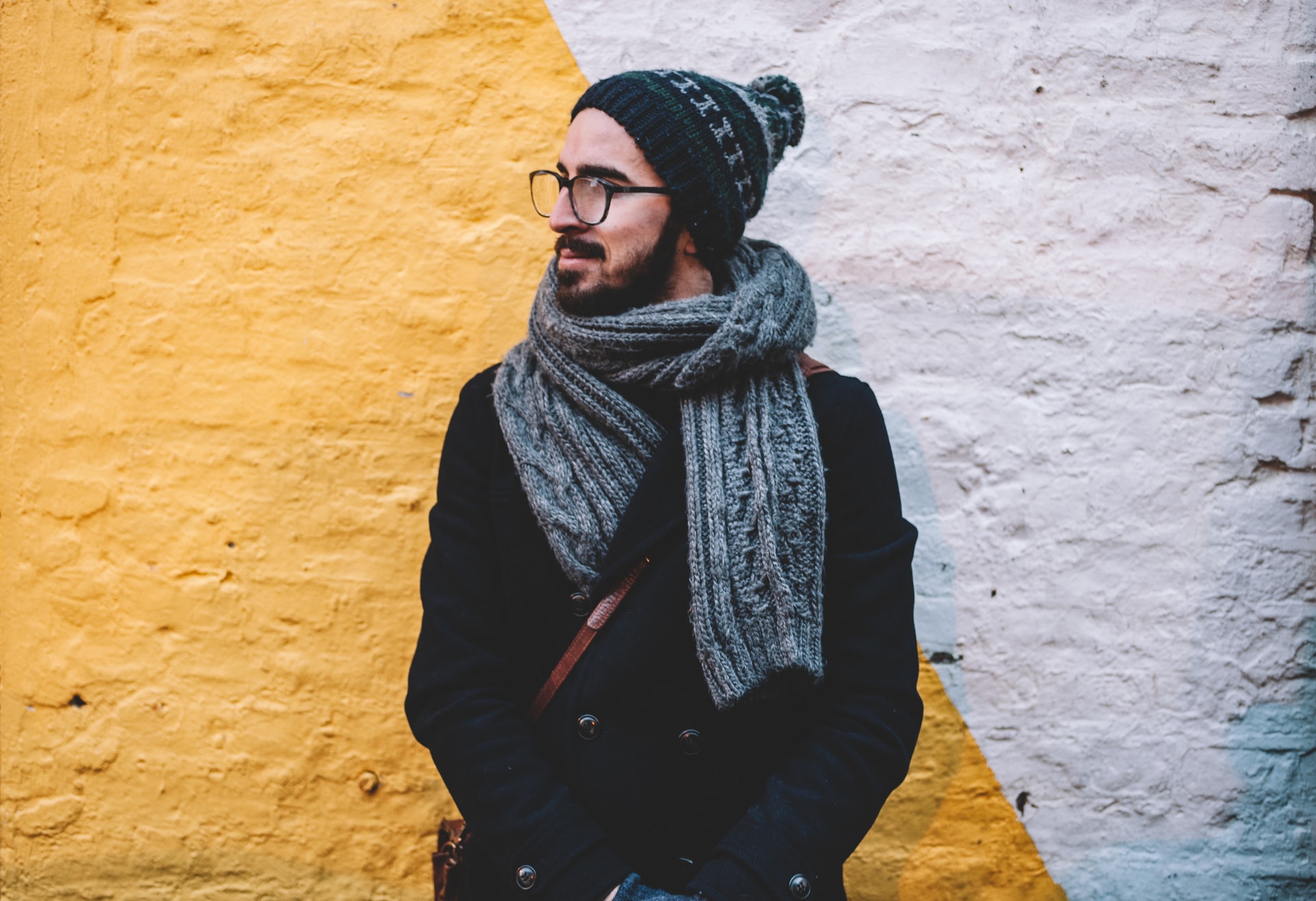 a man looking away from camera with hat and scarf on, leaning against the wall
