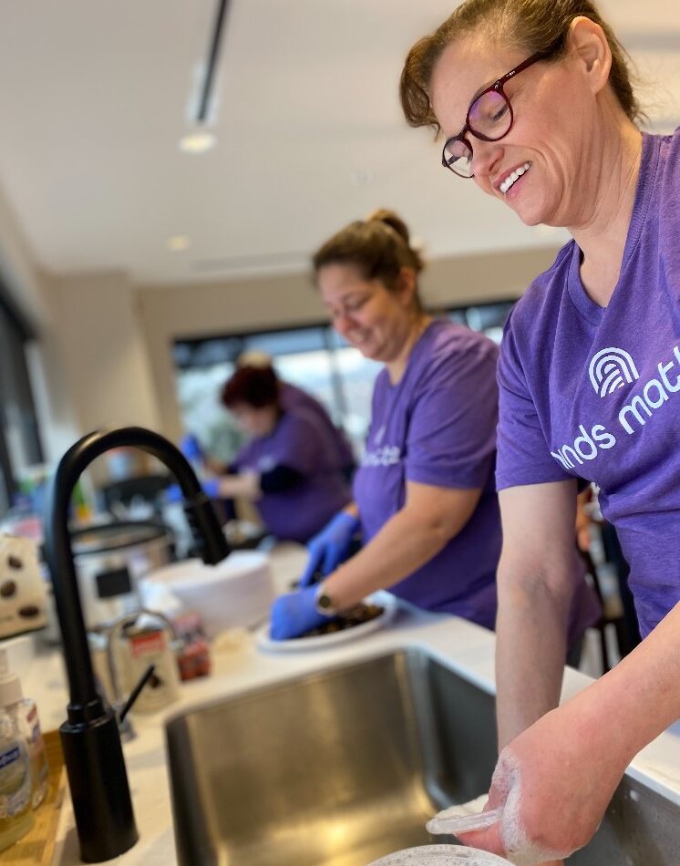 Woman in Minds Matter, LLC purple shirt washing dishes with two other women in purple Minds Matter shirts in background.