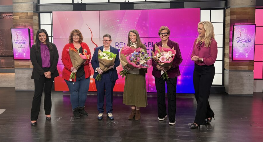 Crystal Martinez, Kristie Morton, Kate Lynch, KK Assmann, Janet Williams, and Abby Eden pose together in front of Remarkable Woman display.