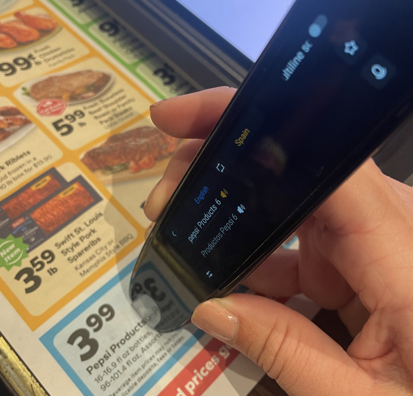A person holds a handheld translation device over a grocery store advertisement. The device is translating English text about Pepsi products into Spanish, displaying both languages on its screen. The ad beneath shows food and beverage promotions with prices, while the translator is positioned directly over a Pepsi products coupon.
