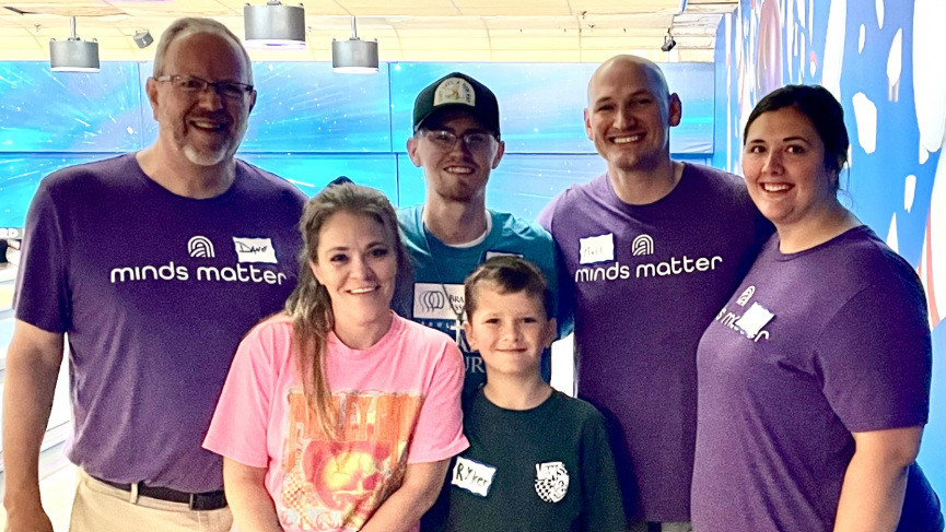 Six participants smile for a group photo at a bowling alley during the Brain Injury Association of Missouri's Bowling for Brain Injury event. Three individuals wearing purple Minds Matter shirts stand alongside a consumer and family members, with bowling lanes and colorful walls visible in the background.